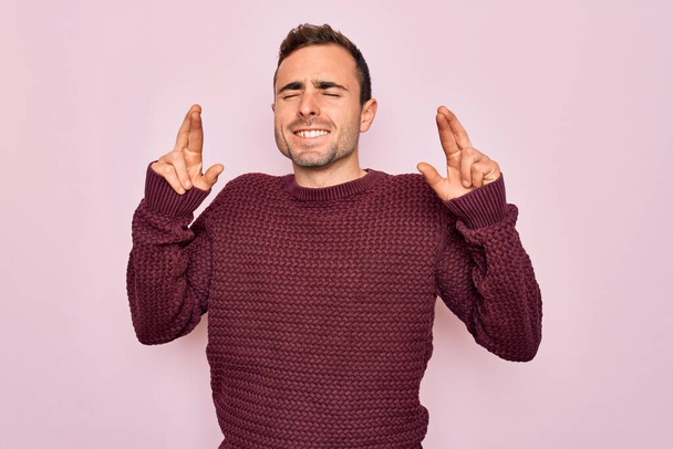Jonge knappe man met blauwe ogen in casual trui die over roze achtergrond staat gebaren vinger gekruist glimlachend met hoop en ogen dicht. Geluk en bijgelovig concept. - Foto, afbeelding