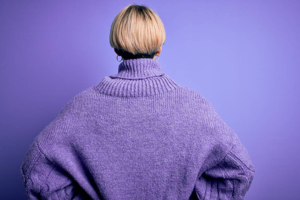 Młoda blondynka z krótkimi włosami nosząca zimowy sweter na fioletowym tle stojąca tyłem do tyłu z rękami na ciele - Zdjęcie, obraz