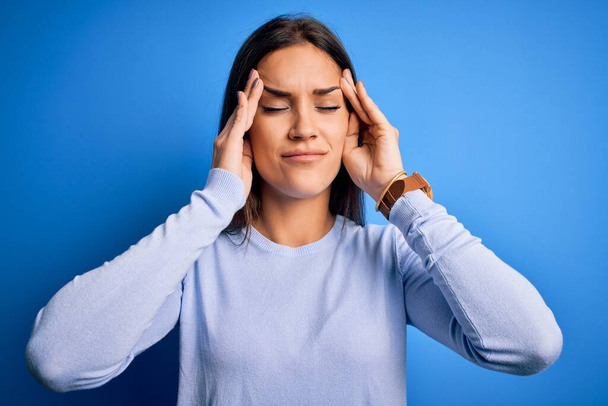 Jeune belle femme brune portant un pull décontracté debout sur fond bleu avec la main sur le mal de tête parce que le stress. Migraine souffrante. - Photo, image