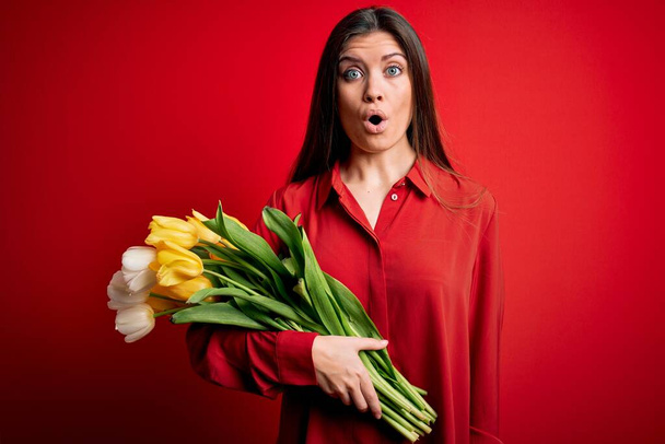 Młoda piękna romantyczna kobieta z niebieskimi oczami trzymająca bukiet pięknych żółtych tulipanów przerażona zaskakującą twarzą, przerażona i podekscytowana ekspresją strachu - Zdjęcie, obraz