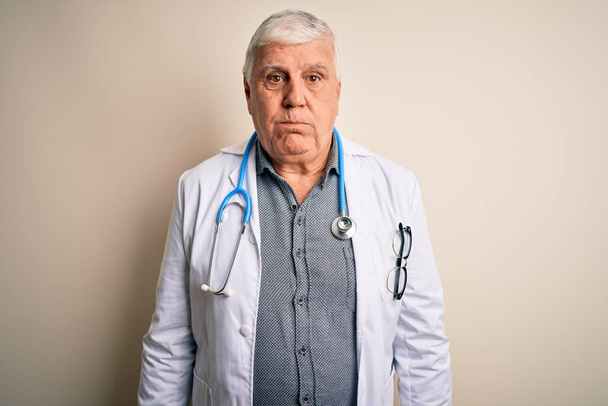 Senior pohledný chraptivý lékař muž v kabátě a stetoskopu přes bílé pozadí nafoukané tváře s legrační tváří. Ústa nadýchaná vzduchem, šílený výraz. - Fotografie, Obrázek