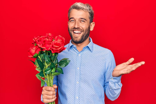 Genç, sarışın, romantik adam kırmızı arka planda gül buketi tutuyor başarıyı mutlu bir gülümsemeyle ve el kaldırarak kutluyor. - Fotoğraf, Görsel