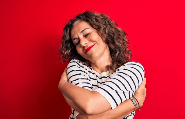Middle age beautiful brunette woman wearing striped t-shirt standing over red background hugging oneself happy and positive, smiling confident. Self love and self care - 写真・画像