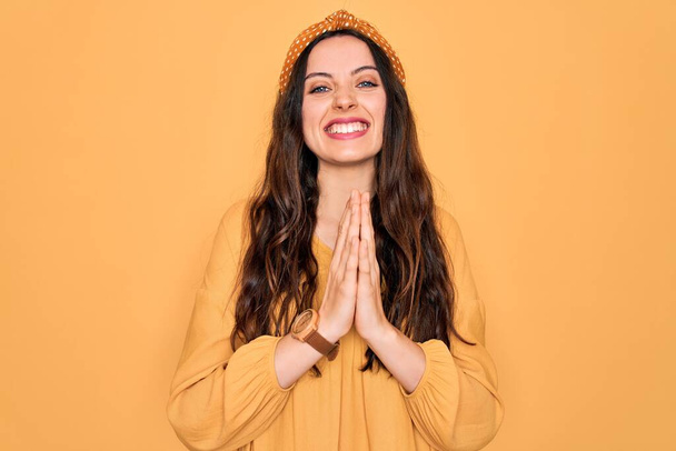 Young beautiful woman wearing casual t-shirt and diadem standing over yellow background praying with hands together asking for forgiveness smiling confident. - Photo, Image