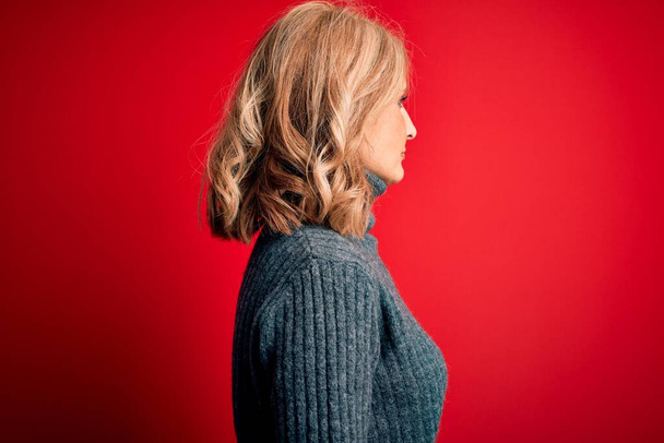Середньовіччя красива блондинка в повсякденному светрі на червоному тлі, дивлячись в бік, розслабляє профільну позу з природним обличчям з впевненою посмішкою
. - Фото, зображення