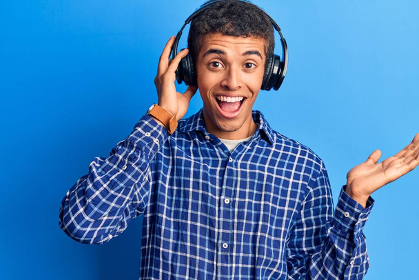 Young african amercian man listening to music using headphones celebrating achievement with happy smile and winner expression with raised hand  - Foto, imagen