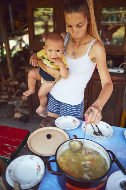 Mujer con bebé preparando la comida y la fiesta para el almuerzo de verano en el campo casa de campo rústica cocina al aire libre
. - Foto, Imagen