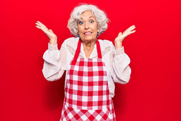 Senior grey-haired woman wearing apron clueless and confused with open arms, no idea and doubtful face.  - Photo, Image