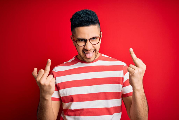 Young handsome man wearing casual striped t-shirt and glasses over isolated red background Showing middle finger doing fuck you bad expression, provocation and rude attitude. Screaming excited - Fotografie, Obrázek