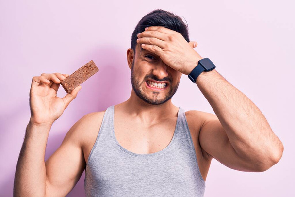 Young handsome man with beard eating energy protein bar over isolated pink background stressed and frustrated with hand on head, surprised and angry face - Fotó, kép