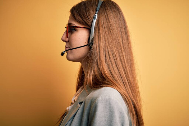 Jovem ruiva agente de call center mulher sobrecarregada usando óculos usando fone de ouvido olhando para o lado, relaxar pose perfil com rosto natural com sorriso confiante
. - Foto, Imagem