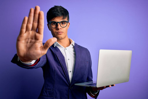 Junger gutaussehender Geschäftsmann mit Brille, der mit Laptop vor lila Hintergrund arbeitet und mit offener Hand Stoppschild mit ernstem und selbstbewusstem Gesichtsausdruck macht, Verteidigungsgeste - Foto, Bild