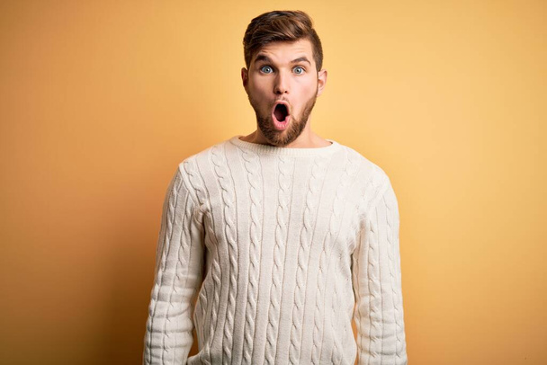 Young blond man with beard and blue eyes wearing white sweater over yellow background afraid and shocked with surprise and amazed expression, fear and excited face. - Foto, Bild