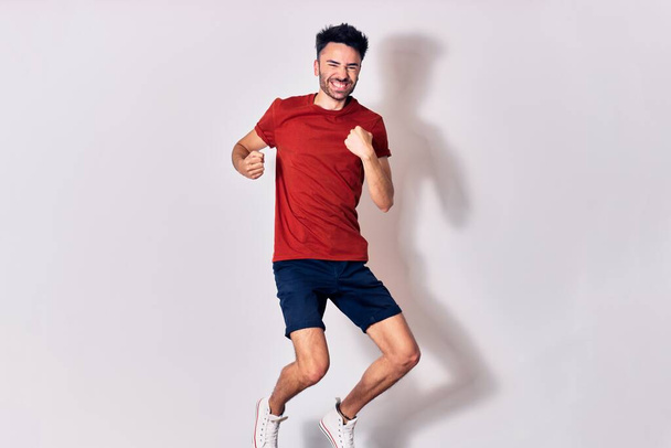Young handsome man wearing casual clothes smiling happy. Jumping with smile on face doing winner gesture with fists up over isolated white background - Φωτογραφία, εικόνα