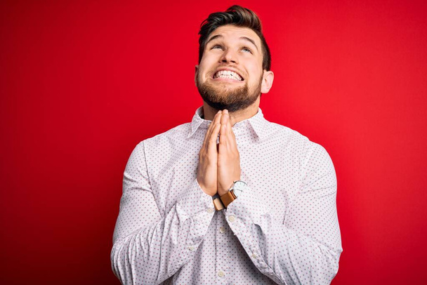 Young blond businessman with beard and blue eyes wearing elegant shirt over red background begging and praying with hands together with hope expression on face very emotional and worried. Begging. - Foto, afbeelding