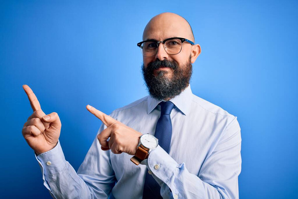 Hombre calvo de negocios guapo con barba con corbata elegante y gafas sobre fondo azul sonriendo y mirando a la cámara apuntando con dos manos y dedos hacia un lado
. - Foto, imagen