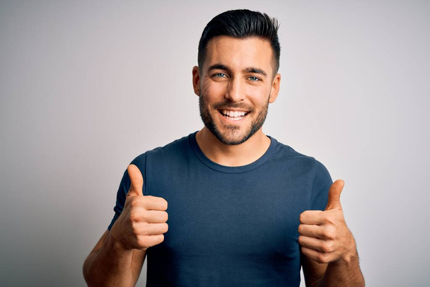 Young handsome man wearing casual t-shirt standing over isolated white background success sign doing positive gesture with hand, thumbs up smiling and happy. Cheerful expression and winner gesture. - Фото, изображение