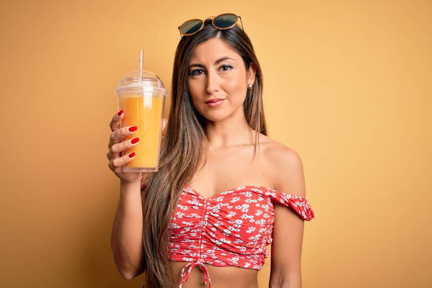 Krásná brunetka žena pití skla se zdravou pomerančovou šťávou přes žluté pozadí s jistým výrazem na chytré tváři myšlení vážné - Fotografie, Obrázek