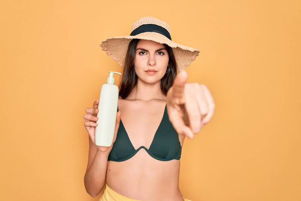 Young beautiful girl wearing swimwear bikini and summer hat holding sun protection cream pointing with finger to the camera and to you, hand sign, positive and confident gesture from the front - Фото, изображение