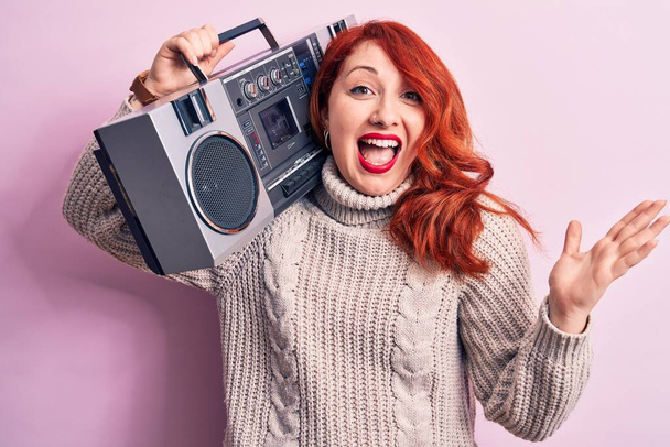 Bella donna rossa che ascolta la musica che tiene il boombox vintage su sfondo rosa che celebra il successo con sorriso felice e l'espressione del vincitore con la mano sollevata - Foto, immagini