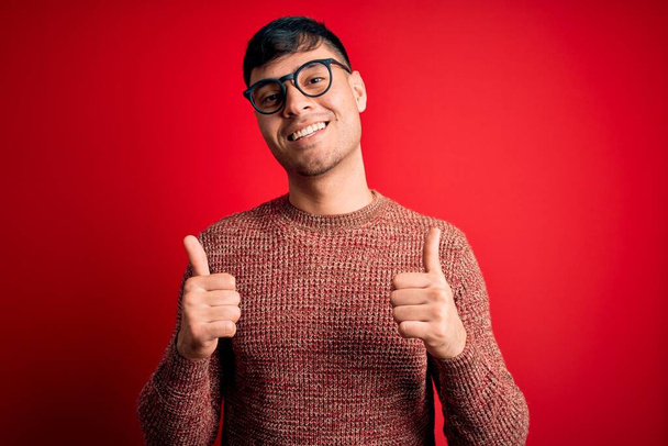 Joven hombre hispano guapo con gafas nerd sobre el signo de éxito de fondo rojo haciendo gesto positivo con la mano, pulgares hacia arriba sonriendo y feliz. Expresión alegre y gesto ganador
. - Foto, Imagen