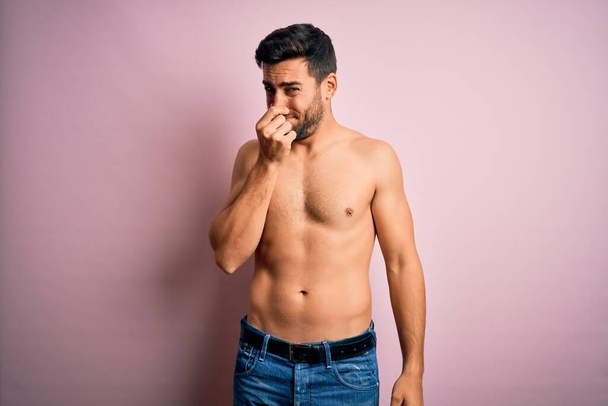 Jonge knappe sterke man met baard shirtloos staan over geïsoleerde roze achtergrond ruiken iets stinkend en walgelijk, ondraaglijke geur, adem inhouden met vingers op de neus. Slechte geur - Foto, afbeelding