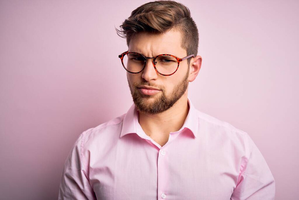 Homem loiro bonito jovem com barba e olhos azuis vestindo camisa rosa e óculos céticos e nervosos, franzindo a testa chateado por causa do problema. Pessoa negativa
. - Foto, Imagem