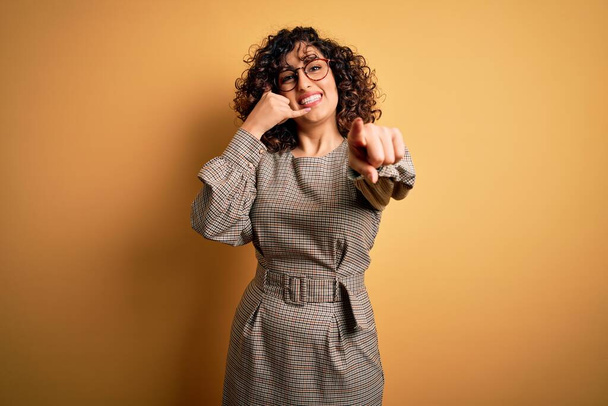 Beautiful arab business woman wearing dress and glasses standing over yellow background smiling doing talking on the telephone gesture and pointing to you. Call me. - Foto, Imagem