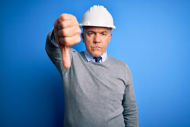 Der gut aussehende grauhaarige Ingenieur mittleren Alters, der einen Schutzhelm über blauem Hintergrund trägt, sieht unglücklich und wütend aus und zeigt Ablehnung und Ablehnung mit Geste nach unten. Schlechte Miene. - Foto, Bild