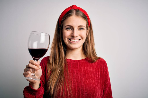 Jeune belle rousse femme buvant un verre de vin rouge sur fond blanc isolé avec un visage heureux debout et souriant avec un sourire confiant montrant des dents - Photo, image
