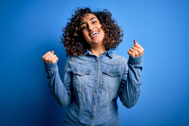 Young beautiful curly arab woman wearing casual denim shirt standing over blue background celebrating surprised and amazed for success with arms raised and open eyes. Winner concept. - Foto, imagen