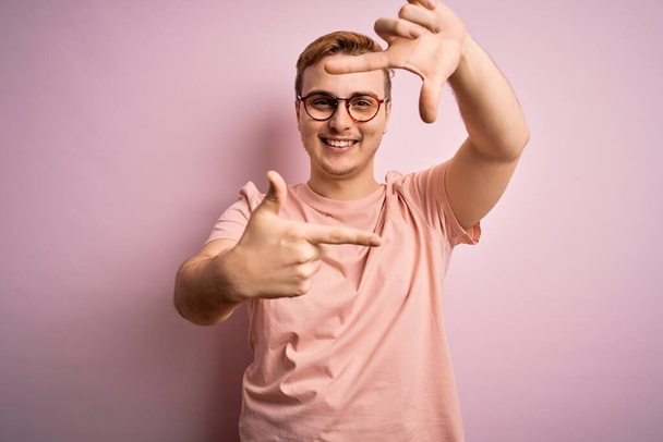 Young handsome redhead man wearing casual t-shirt standing over isolated pink background smiling making frame with hands and fingers with happy face. Creativity and photography concept. - Photo, Image