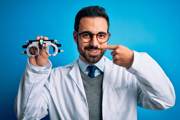 Young handsome optical man with beard holding optometry glasses over blue background Pointing with hand finger to face and nose, smiling cheerful. Beauty concept - Photo, Image
