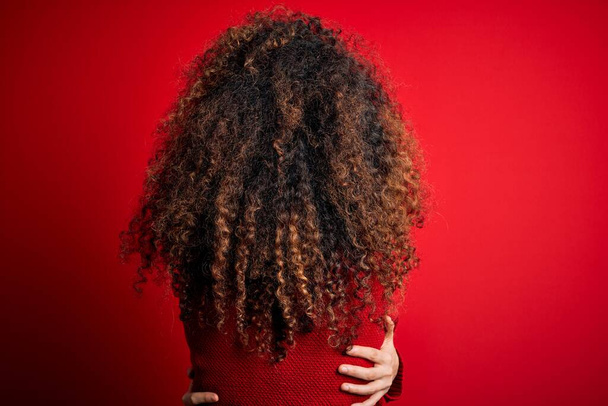 Young beautiful woman with curly hair and piercing wearing casual red sweater Hugging oneself happy and positive from backwards. Self love and self care - Фото, изображение