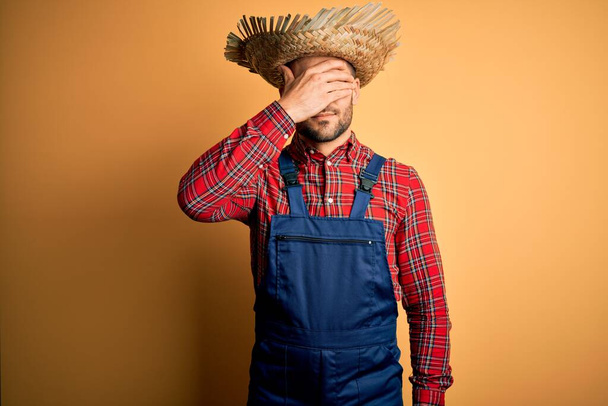 Nuori maaseudun viljelijä mies yllään ruokalappu yleistä ja maaseudun hattu keltainen tausta hymyilee ja nauraa käsin kasvot peittävät silmät yllätys. Sokea käsite. - Valokuva, kuva