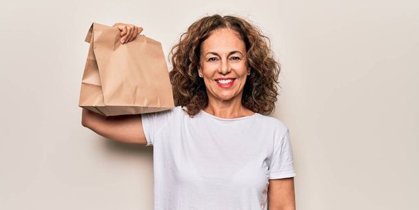 Middle age woman holding deliver paper bag with takeaway food over white background looking positive and happy standing and smiling with a confident smile showing teeth - Photo, image