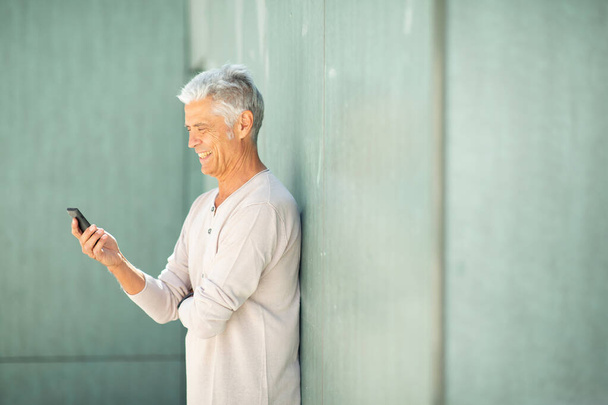 Ein älterer, gutaussehender Mann lehnt an der Wand und blickt auf sein Handy - Foto, Bild