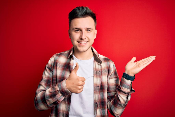 Fiatal jóképű kaukázusi férfi visel alkalmi modern ing felett piros elszigetelt háttér Mutogató tenyér kéz és csinál oké gesztus hüvelykujjal fel, mosolygós boldog és vidám - Fotó, kép