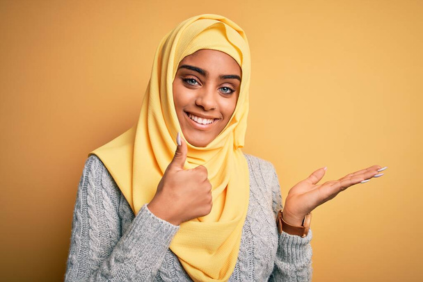 Νεαρή όμορφη Αφροαμερικανή κοπέλα που φοράει muslim hijab πάνω από απομονωμένο κίτρινο φόντο Εμφανίζει το χέρι παλάμη και κάνει ok χειρονομία με τους αντίχειρες επάνω, χαμογελώντας χαρούμενος και χαρούμενος - Φωτογραφία, εικόνα