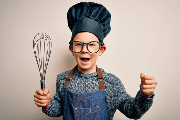 Young little caucasian cook kid wearing chef uniform and hat using manual whisk screaming proud and celebrating victory and success very excited, cheering emotion - Photo, Image