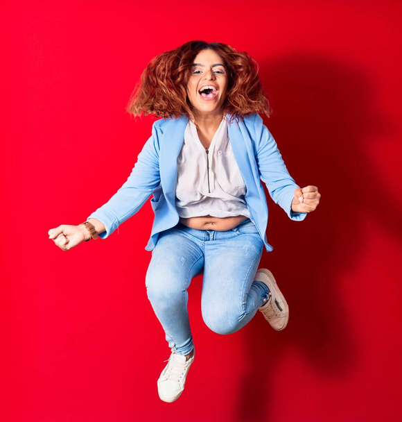 Young beautiful latin woman wearing casual clothes smiling happy. Jumping with smile on face doing winner gesture with fists up over isolated red background - Fotografie, Obrázek