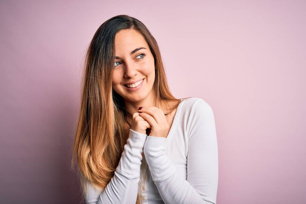 Young beautiful blonde woman with blue eyes wearing white t-shirt over pink background laughing nervous and excited with hands on chin looking to the side - Fotó, kép