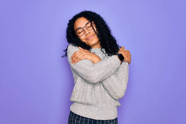 Nuori afrikkalainen amerikkalainen nainen yllään rento villapaita ja lasit violetti tausta halaamalla itseään onnellinen ja positiivinen, hymyillen luottavainen. Itserakkaus ja itsehoito - Valokuva, kuva