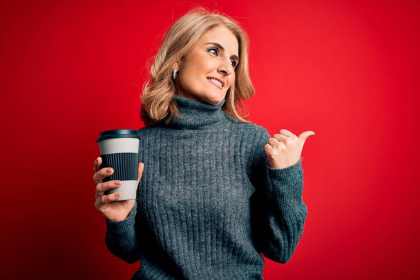 Edad media hermosa mujer rubia bebiendo una taza de café para llevar sobre fondo rojo señalando y mostrando con el pulgar hacia el lado con la cara feliz sonriendo
 - Foto, Imagen