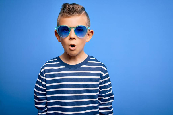 Young little caucasian kid with blue eyes standing wearing sunglasses over blue background afraid and shocked with surprise expression, fear and excited face. - Photo, Image