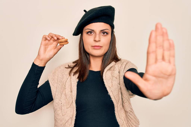 Młoda piękna brunetka kobieta nosząca beret jedząc francuski makaron na białym tle z otwartą ręką robi znak stop z poważnym i pewnym siebie wyrazem twarzy, gest obrony - Zdjęcie, obraz