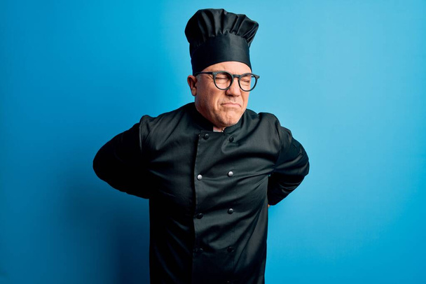 Středního věku pohledný šedovlasý kuchař v kuchařské uniformě a klobouku Trpí bolestí zad, dotýká se rukou, svalová bolest - Fotografie, Obrázek