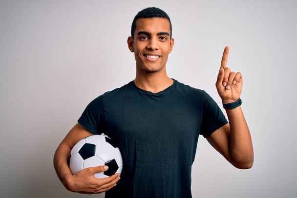 Όμορφος Αφροαμερικάνος που παίζει ποδόσφαιρο κρατώντας μπάλα πάνω από λευκό φόντο με ένα μεγάλο χαμόγελο στο πρόσωπο, δείχνοντας με το χέρι το δάχτυλο στο πλάι κοιτάζοντας την κάμερα. - Φωτογραφία, εικόνα