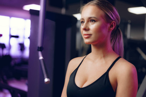 Close up portrait of a young blonde woman in sport bra in a dark gym - Fotó, kép