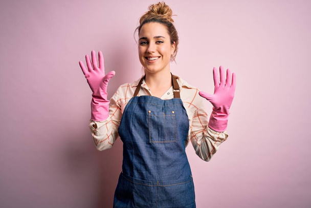 Joven hermosa mujer rubia limpiadora haciendo tareas domésticas con arpon y guantes mostrando y señalando con los dedos número diez mientras sonríe confiado y feliz
. - Foto, imagen